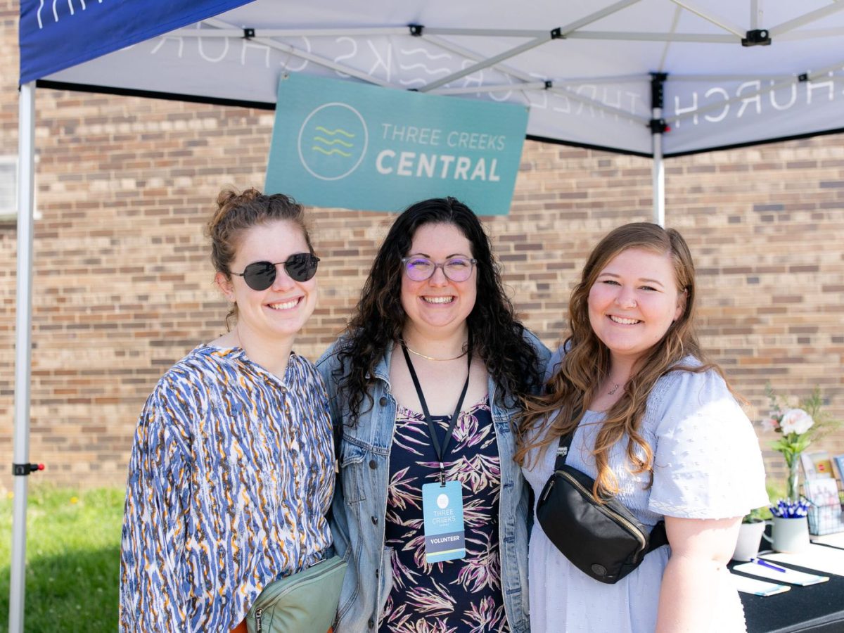 Three volunteers smiling under the Three Creeks Church canopy at the Three Creeks Central welcome table.