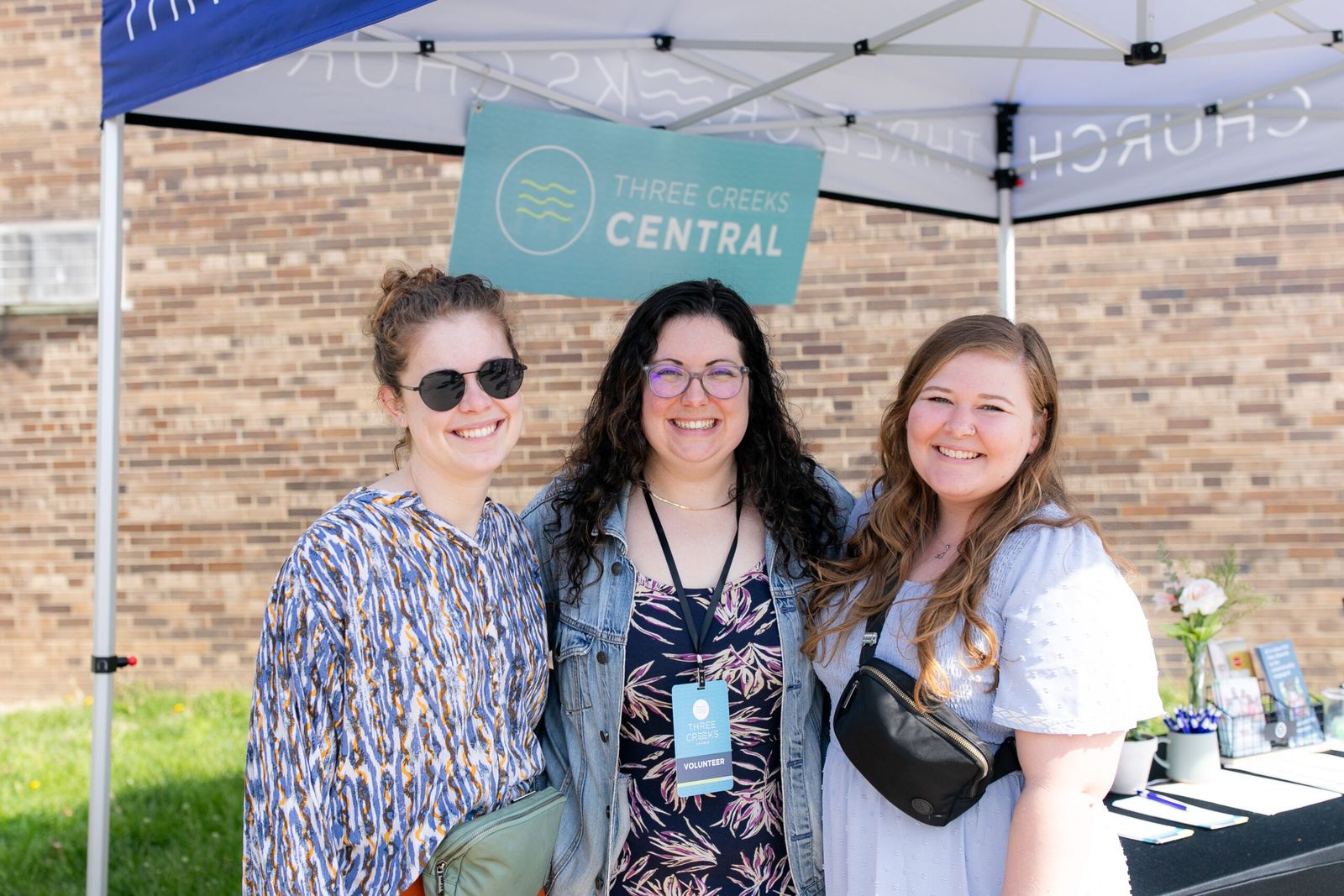 Three volunteers smiling under the Three Creeks Church canopy at the welcome table.
